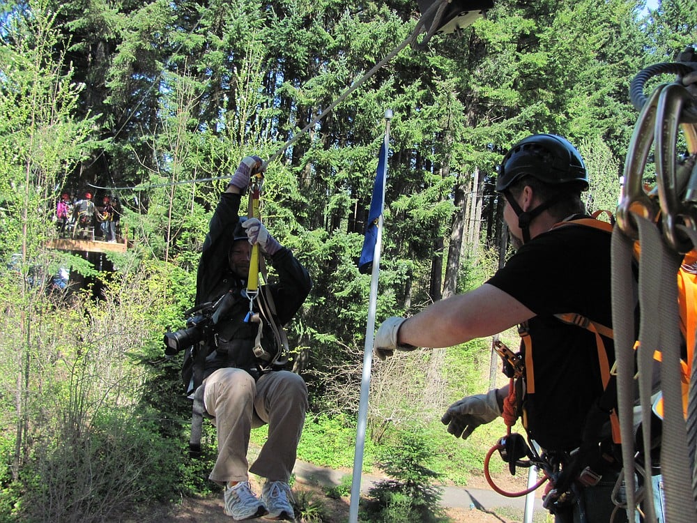 Guide Shayne Large assists Troy Wayrynen, a photographer with The Columbian, on the first "zip" of the tour, a 100 foot long   inch galvanized aircraft cable line.