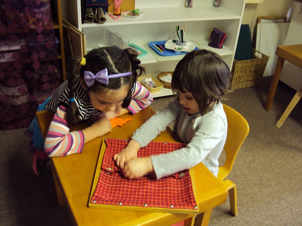Lila McCarthy, 5, (left) leans in to help fellow Montessori student Adalie Hillyard, 3, with a button frame.