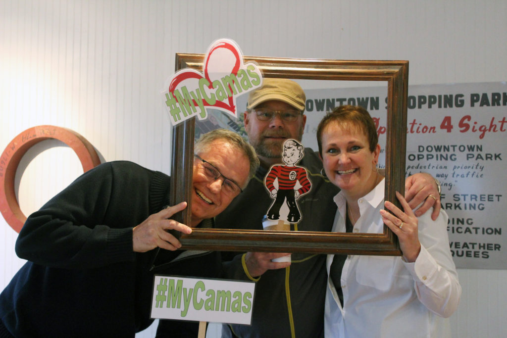 Downtown Camas Association Executive Director Carrie Schulstad (right) poses inside the #MyCamas frame at the DCA office with Camas Parks and Recreation Board Chair Randy Curtis (left) and Windermere real estate broker Greg Goforth (center) Tuesday morning, before finding out Camas had made the top 10 cities in the running for the fourth season of the online show, 