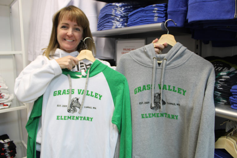 Sheryl Obegi, one of the co-owners of the new Papermaker Pride business in downtown Camas, shows the store&#039;s Grass Valley Elementary-themed shirts. In addition to being a business owner, Obegi works as a third-grade teacher at Grass Valley.