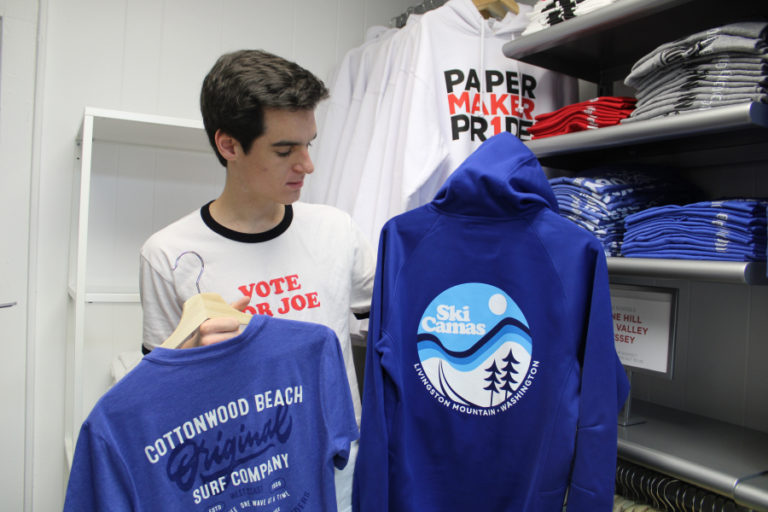 Camas High School senior Tommy Kelly holds up two Camas-themed shirts featuring Livingston Mountain and Cottonwood Beach, at the newly opened Papermaker Pride shop in downtown Camas.