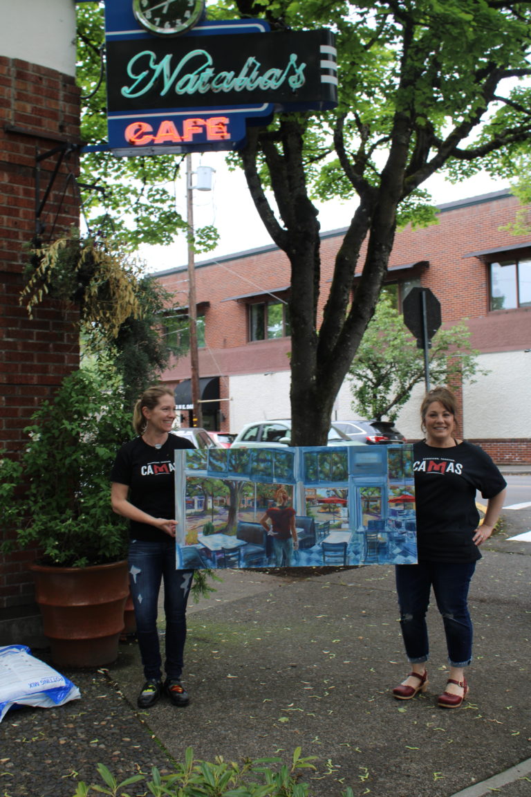 (Kelly Moyer/Post-Record) Natalia's Cafe owner Erica Slothower (right) and manager Wendy DelBosque (left) hold Camas painter Anna Norris' recent work, "We're Not in Camas Anymore," featuring DelBosque looking out the cafe windows onto the desolate streets of downtown Camas during the height of the COVID-19 quarantine, on Friday, May 22.