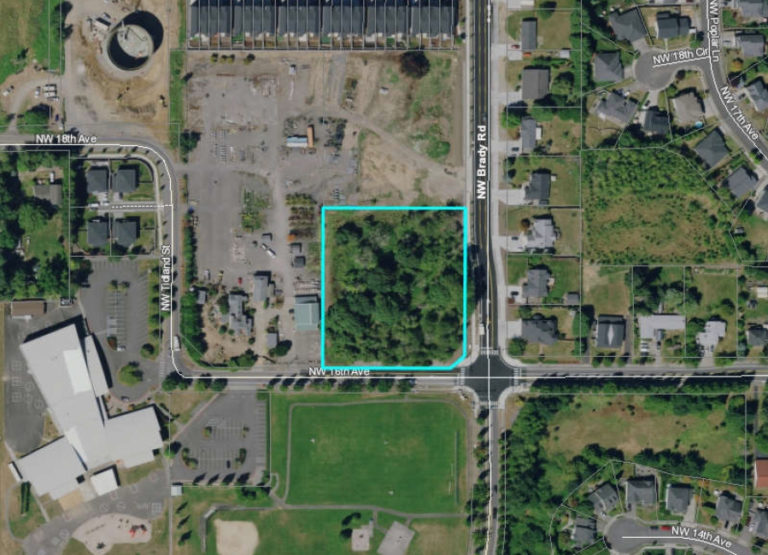 An aerial photo shows a vacant 2.16-acre parcel zoned for community commercial use and slated for a proposed gas station-convenience store development (outlined in blue), located at the intersection of Northwest Brady Road and Northwest 16th Avenue, across from the Prune Hill Sports Park (green space below vacant parcel) and down the street from the Prune Hill Elementary School (white roof, lower left corner).