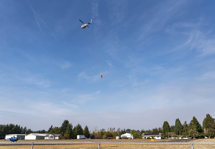 Smoke from the Nakia Creek Fire is visible on the horizon as a helicopter carrying a water bucket flies over Grove Field north of Camas on Monday, Sept. 17, 2022. (Contributed photo courtesy of Taylor Balkom/The Columbian)