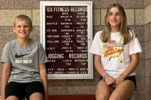 Brady Marshall, left, and his older sister, Eliisa, sit in front of Dorothy Fox Elementary School's fitness leaderboard in September 2023. The siblings finished the mile run in 5 minutes, 45 seconds, as fifth-graders, tying for the school's record. (Contributed photo courtesy of Cathy Sork)